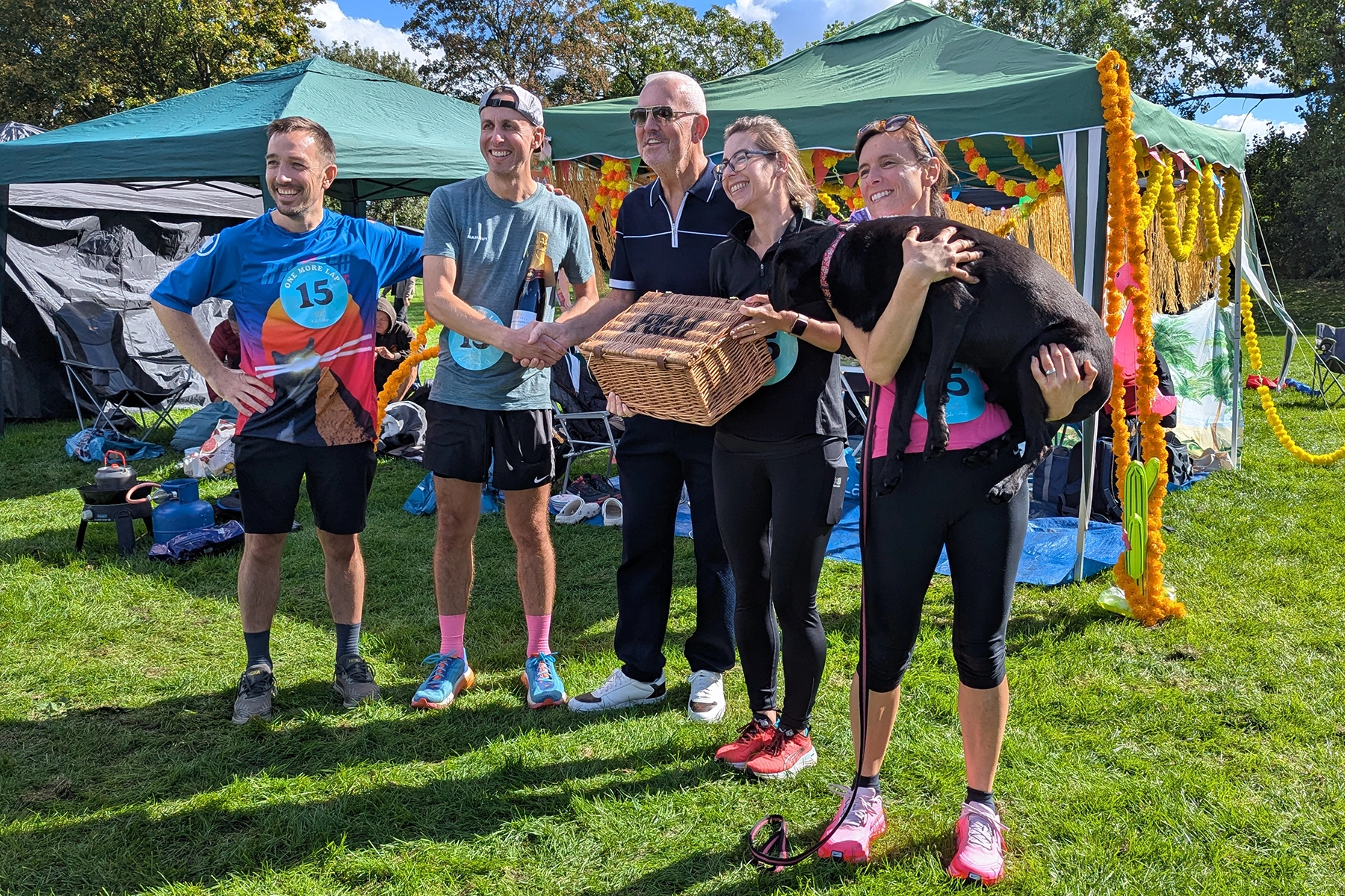 Man wearing a black and red running outfit with a race bib numbered 2 smiles while jogging on a park path, followed by a group of runners in brightly colored shirts, surrounded by lush greenery.