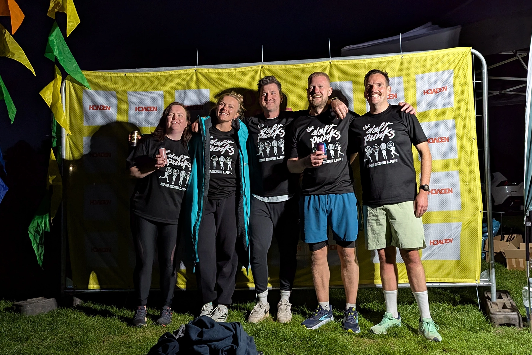Man wearing a black and red running outfit with a race bib numbered 2 smiles while jogging on a park path, followed by a group of runners in brightly colored shirts, surrounded by lush greenery.